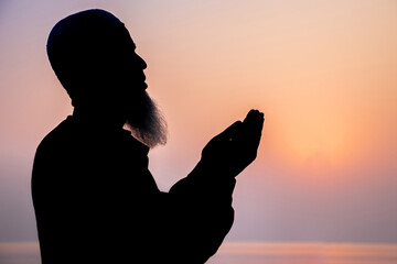 Asian Muslim bearded man praying with hands raised to God on sunset background. Silhouette of an...