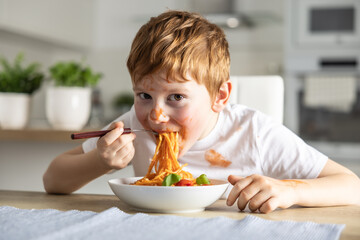 A cute little boy is eating spaghetti bolognese for lunch in the kitchen at home and is covered in ketchup