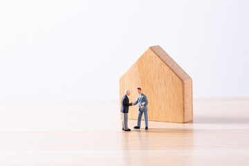木製の住宅模型の前で握手するミニチュアビジネスマン