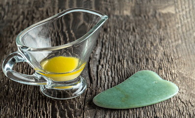 Jade scraper for gua cha and melted ghee in a glass cup on a background of wood texture.