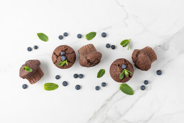 Fresh baked muffins with chocolate chips, blueberry berries and mint leaves on white marble table background.