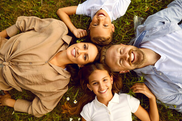Close up top view of happy family of four lying in circle on green grass and looking at camera in summer park. Smiling parents having fun with kids boy and girl in nature enjoying time together.