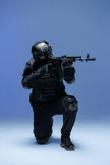 Soldier in black military uniform for city with helmet, action camera and machine gun, photo in studio. Army man stands on one knee, defensive position