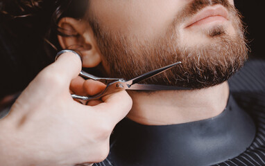 Hipster man sitting in armchair barber shop while hairdresser shaves beard with scissors