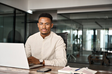 Fototapeta na wymiar Confident young professional African American business man looking at camera sitting at work desk using computer. Portrait of male company employee or entrepreneur with laptop in office, copy space.