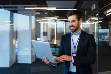 Fototapeta premium Close-up photo of a smiling young businessman. a specialist programmer standing in a suit and with a badge in the office at a conference, holding and using a laptop.