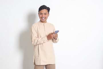 Portrait of young excited Asian muslim man in koko shirt holding mobile phone with smiling expression on face. Social media and internet concept. Isolated image on white background