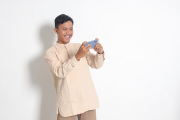 Portrait of young excited Asian muslim man in koko shirt holding mobile phone and playing games on his smartphone. Isolated image on white background