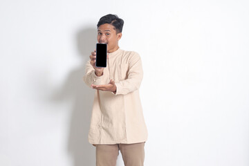 Portrait of young excited Asian muslim man in koko shirt showing blank screen mobile phone mockup while pointing and presenting product. Social media concept. Isolated image on white background