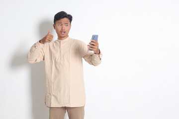 Portrait of young excited Asian muslim man in koko shirt holding mobile phone, taking picture of himself or selfie, saying hi and waving his hand. Social media concept. Isolated on white background