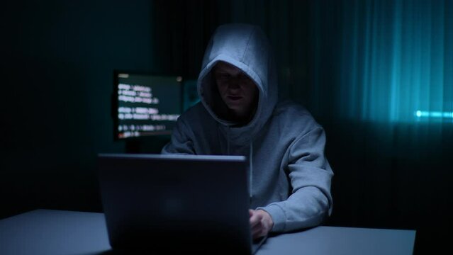 Front view of stealthy hacker man wearing sweatshirt with hood typing on keyboard breaking password using laptop, sitting at desk in dark room, computer monitor with text code on screen on background.
