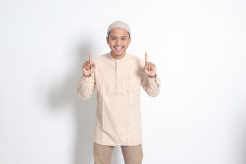 Portrait of shocked Asian muslim man in koko shirt with skullcap showing product and pointing with...