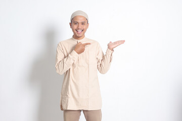 Portrait of shocked Asian muslim man in koko shirt with skullcap showing product and pointing with his hand and finger to the side. Advertising concept. Isolated image on white background