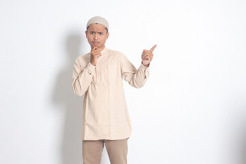 Portrait of shocked Asian muslim man in koko shirt with skullcap showing product and pointing with...