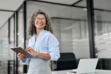 Smiling mature professional business woman bank manager, older happy female executive or lady entrepreneur holding digital tablet pad standing in office at work, looking away at copy space. - Powered by Adobe