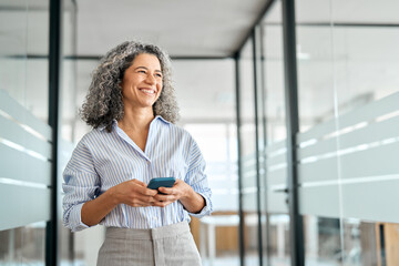 Happy middle aged business woman holding mobile cell phone using cellphone in office. Smiling mature old professional lady business investor owner entrepreneur using smartphone standing looking away. - obrazy, fototapety, plakaty