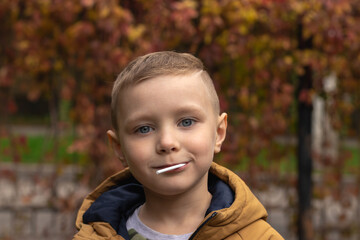 Portrait of a 6 years old boy in light brown coat with a lollipop in his mouth in a autumn park