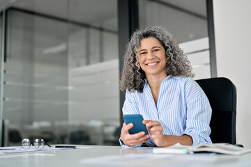 Happy middle aged senior curly business woman holding phone in office looking at camera. Smiling...