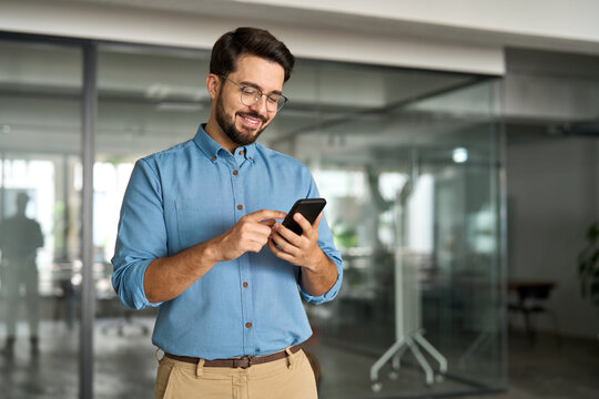 Happy young latin business man holding smartphone standing in office. Smiling hispanic businessman entrepreneur or manager using financial banking apps on cell phone technology device at work.