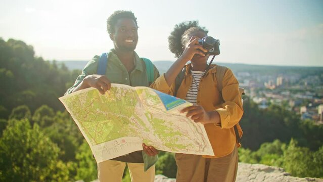 Close view of African American man and woman holding paper map together. Learning topography. Woman making photo with old professional camera. Sightseeing together with beloved one.