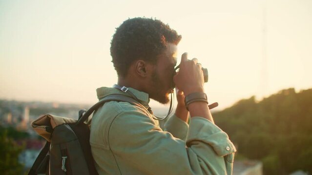 Side view of African American male with beard making photo with his camera. Bearded man with backpack and green t-shirt exploring nature. Making video or professional photos. Concept of photography.
