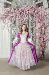 Obraz na płótnie Canvas Beautiful woman in fantasy white and purple rococo style medieval dress standing near wall with pink flowers