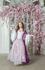 Obraz na płótnie Canvas Beautiful woman in fantasy white and purple rococo style medieval dress standing near wall with pink flowers