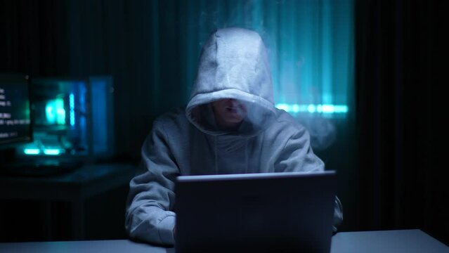 Unrecognizable hacker man in hoodie vaping electronic cigarettes and hacking online web site or engaging hacking into security systems at dark room. Hooded freelancer working laptop computer from home
