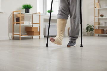 Close up photo of a handicapped man with broken leg trying to walk using his crutches at home. Disabled guy with plastered leg in rehabilitation and recovery having fractured limb. - obrazy, fototapety, plakaty