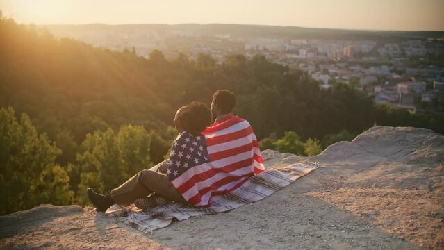 Camera view from behind of African American couple. People sitting on blanket on edge of mountain and covering themselves with American national flag. Observing sunset together in front of them.