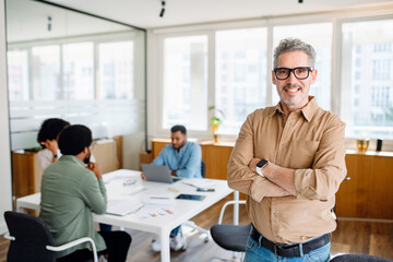A senior professional with arms folded stands confidently in a bright office, as his team engages in focused work behind him, exemplifying a leader overseeing a productive team