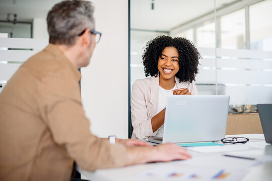 A joyful female professional with curly hair shares a warm smile while engaging in a productive conversation with her mature male colleague in office, depicting an atmosphere of amicable collaboration