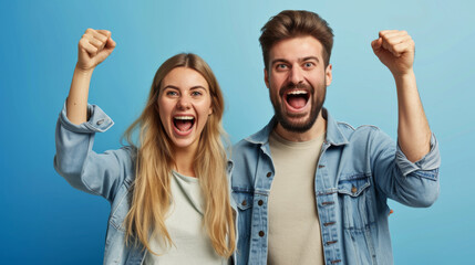 man and a woman are both cheering excitedly with their fists raised, wearing denim jackets, against a blue background
