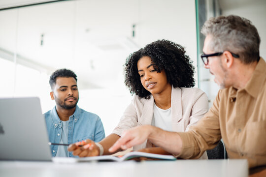 Focused businesswoman in chic blazer leans over to discuss a document with her colleagues, demonstrating teamwork and active problem-solving in a contemporary office. Work-team solving tasks together