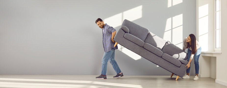 Family Couple Moving Furniture At Home. Young Man And Woman Bought Sofa And Are Placing It In Living Room Together. Two Happy People Moving Gray Couch Inside New House Or Apartment. Banner Background