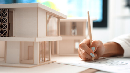 Closeup image of young beautiful caucasian architect engineer hand write mistake points on blueprint with house model placed on table at office. Creative and professional design concept. Immaculate.