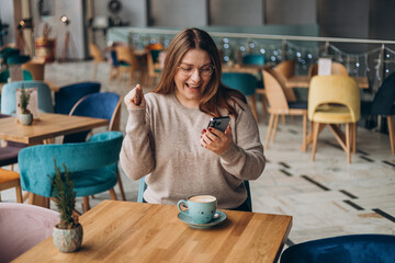 Satisfied young woman with coffee cup holding phone and celebrating in a cafe. hipster female in glasses overjoyed with winning online contest on web site - obrazy, fototapety, plakaty