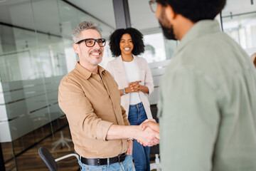 A smiling man in a tan shirt firmly shakes hands with a colleague in a green shirt, symbolizing a successful agreement or introduction, showcasing a friendly and collaborative office environment