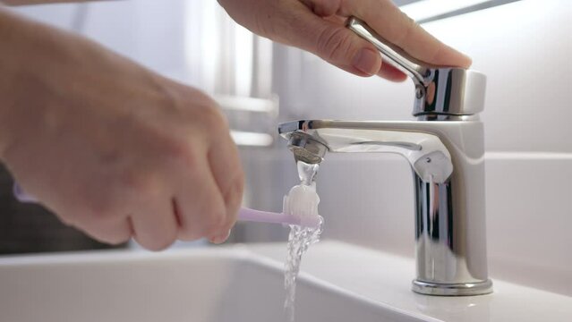 purple toothbrush is rinsed under flowing water from shiny tap in white, well-lit bathroom.