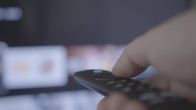 Man watches smart TV and uses black remote control. Person controls TV using a modern remote control choosing channels from a smart tv. Closeup 4k cinematic shot. Shallow depth of field