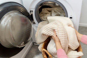 Woman putting white wool sweater and clothes into the drum of a washing machine in laundry room