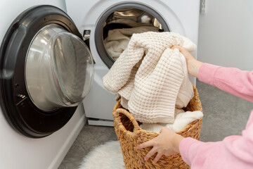 Woman putting white wool sweater into the drum of a washing machine in laundry room