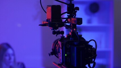 Camera on a tripod against a blurred background of a female presenter in a dark studio in pink, blue neon lights.
