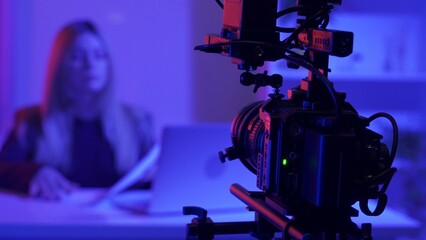 Professional video camera on a tripod against a blurred background of a female presenter in a dark...