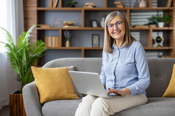 Portrait of senior gray-haired woman at home. A woman is sitting on a sofa in the living room, smiling and looking at the camera, working with a laptop remotely.