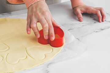 The hands of a caucasian teenage girl cuts out a shortbread dough with a red plastic heart shape .