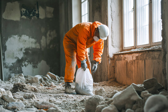 Builder cleaning room after demolition jobs, preparing for room renovation, collecting rubble, wearing gloves, boots and orange coveralls