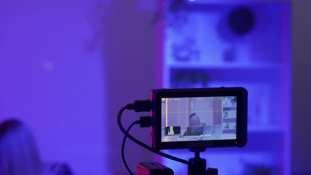 Camera on a tripod against a blurred background of a female presenter in a dark studio in pink, blue neon lights. The camera is lowered, the zoom is adjusted, and the image of the presenter is focused