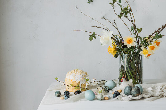 Happy Easter! Stylish easter eggs, homemade easter bread and spring flowers on linen napkin on rustic table against wall. Natural painted blue and marble eggs and daffodils bouquet still life