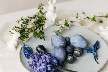 Fototapeta na wymiar Stylish easter eggs on vintage plate, bunny and spring flowers on rustic table. Happy Easter! Natural dye blue eggs, purple hiacynt blossoms on linen napkin. Holiday setting, minimal still life